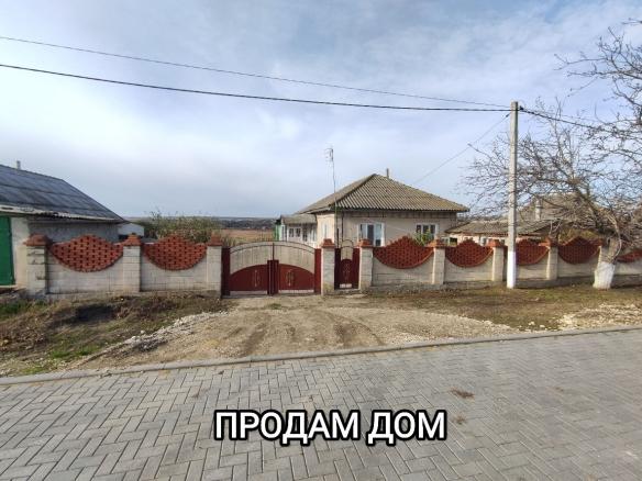 Одноэтажный загородный дом за декоративным бетонным забором с воротами, грунтовым въездом, мощёной улицей и пасмурным небом.