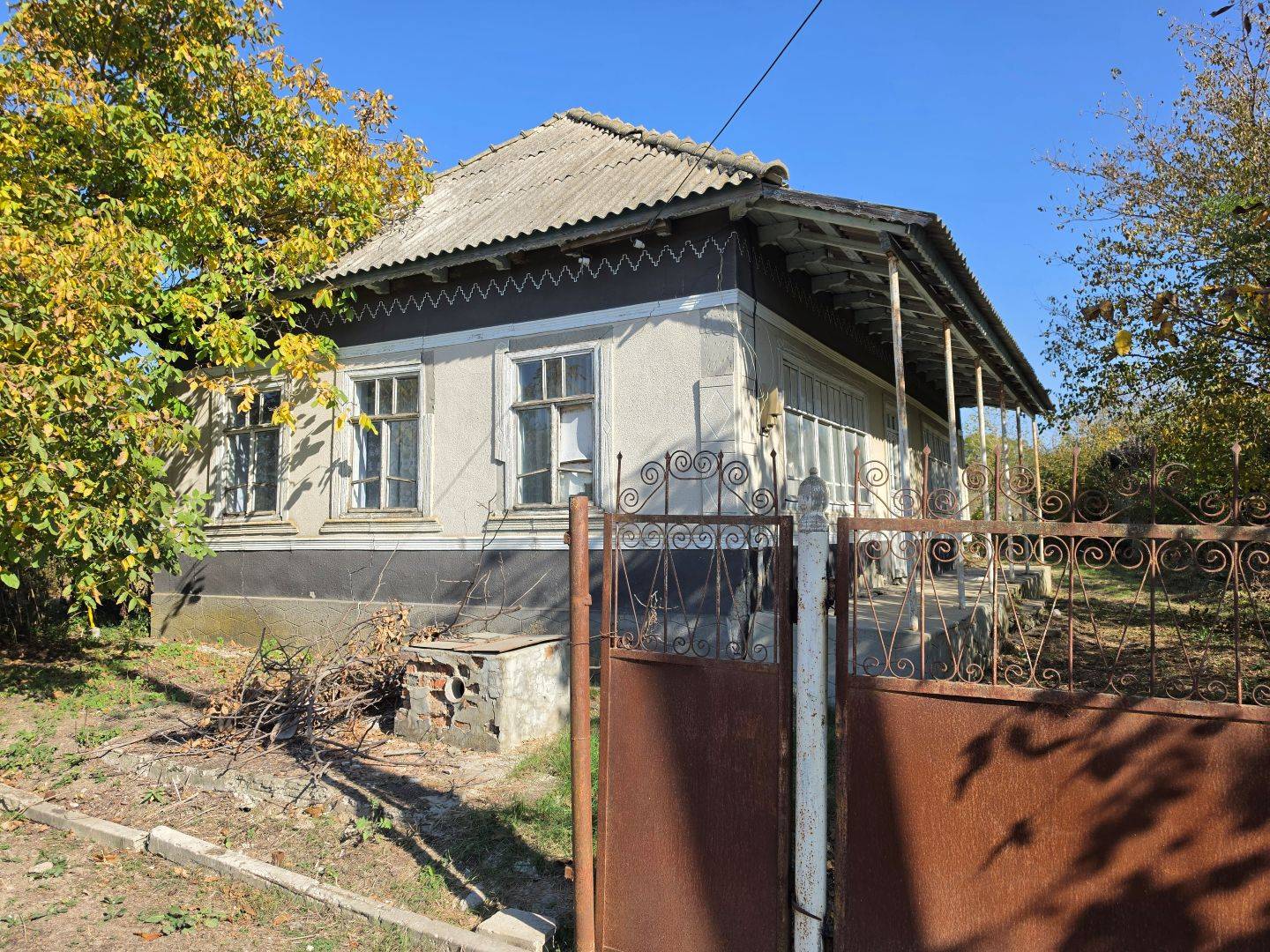 Традиционный сельский дом с деревянными окнами, декоративной отделкой фасада и старинными коваными воротами. Осенние деревья во дворе. Село Кириет-Лунга, Гагаузия
